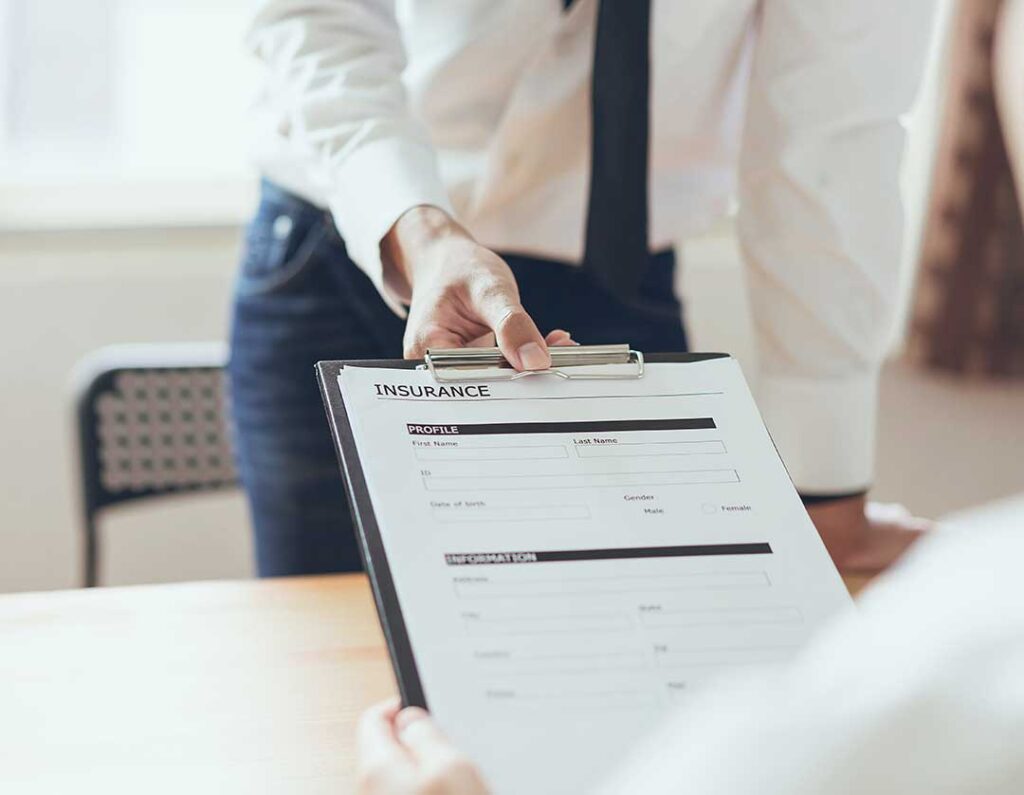 A couple reviews home and auto insurance documents beside a laptop and vehicle keys at a kitchen table.